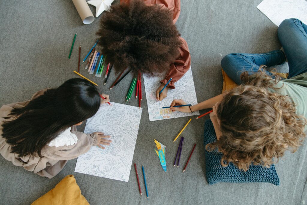 Three diverse young people sitting on the floor together, coloring intricate drawings with colored pencils spread around them. 