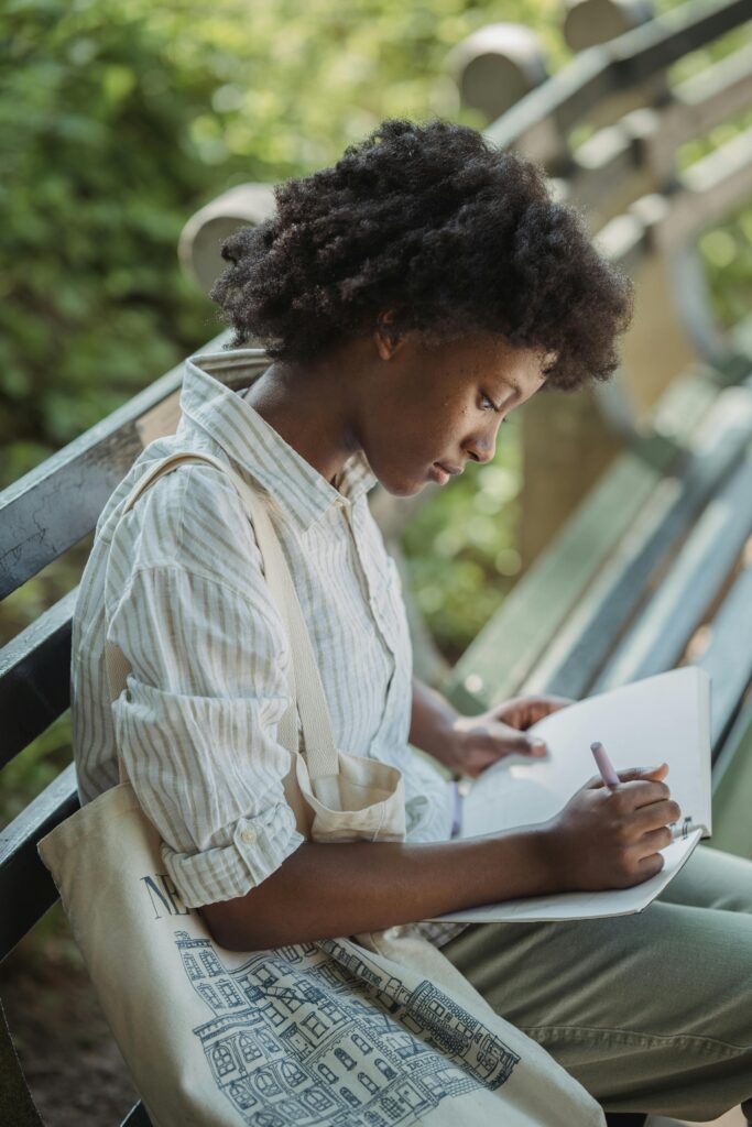 A young woman sitting on an outdoor bench writing in a notebook, surrounded by greenery, representing journaling for mental health and personal growth.