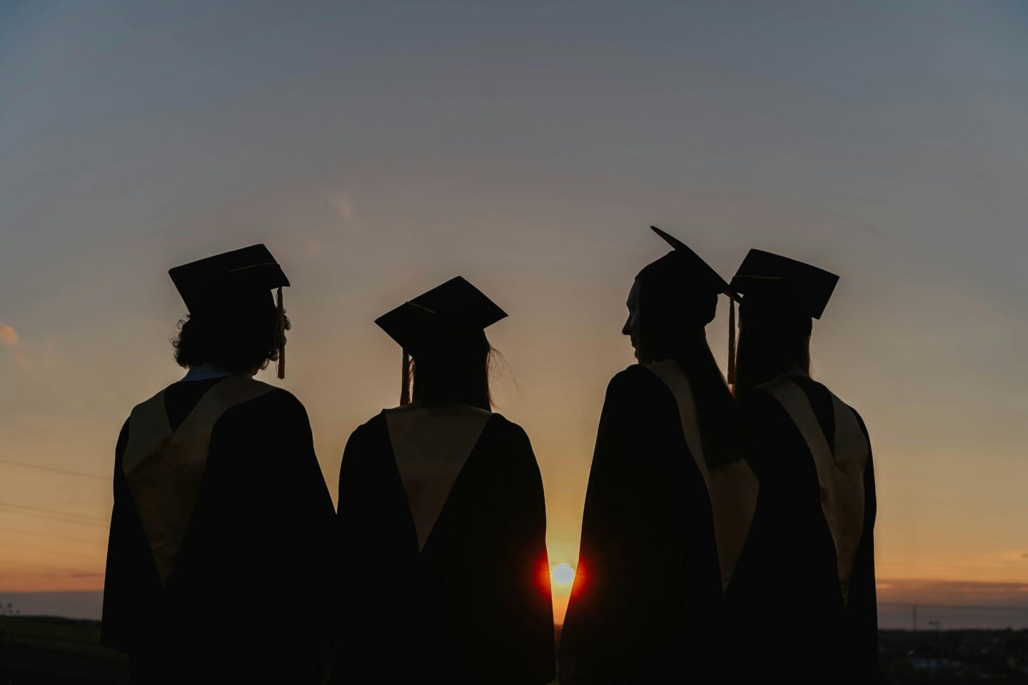 Four graduates in cap and gown standing together as silhouettes against a sunset, representing young adults stepping into a new chapter of life.