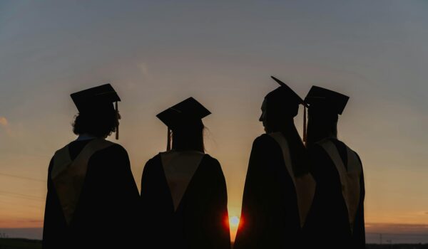 Four graduates in cap and gown standing together as silhouettes against a sunset, representing young adults stepping into a new chapter of life.