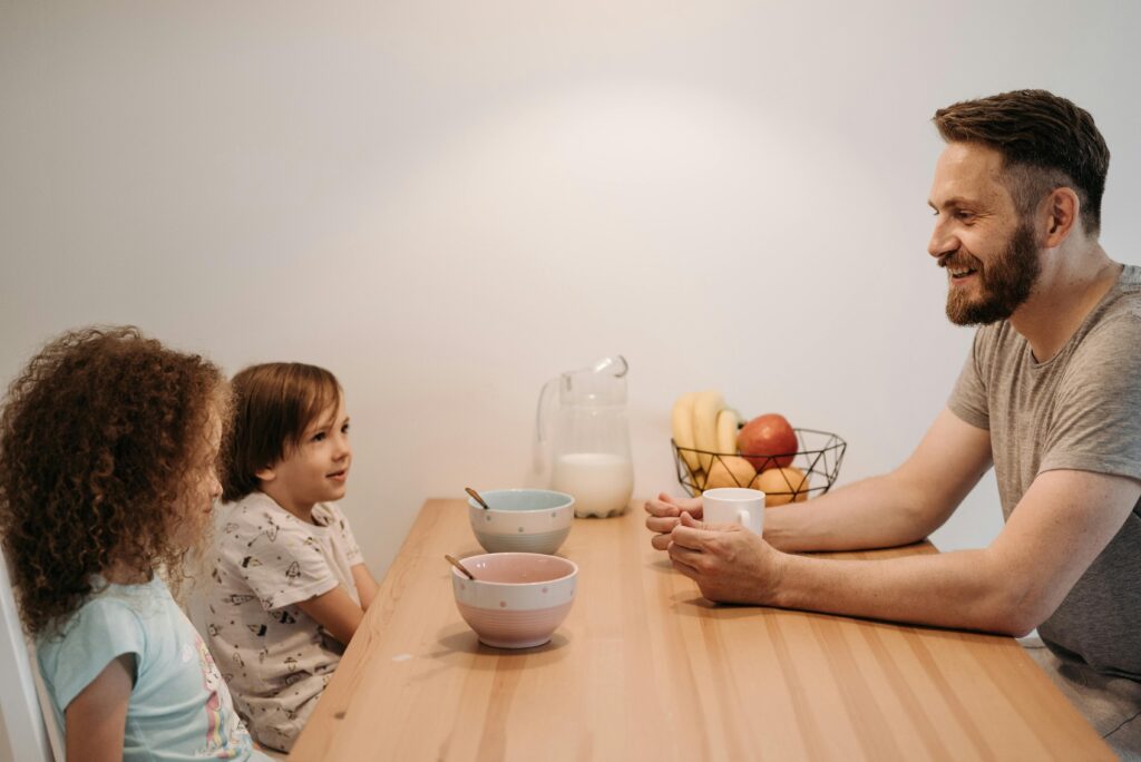 A father smiling and talking with his two young children at a kitchen table during breakfast, representing open and low-pressure family communication.