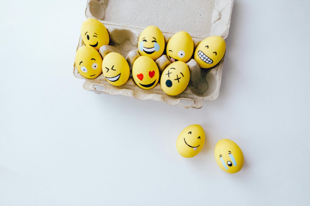 Yellow eggs painted with various emoji expressions including happy, laughing, and sad faces, arranged in a cardboard egg carton — representing the range of emotions children may experience