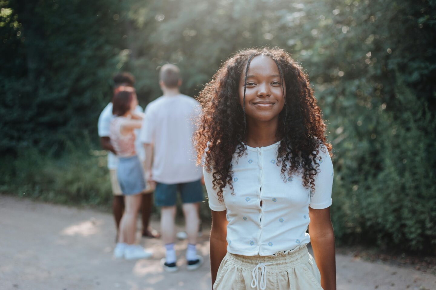 A teenage girl smiling confidently outdoors on a sunny day with friends in the background, representing presence and connection.