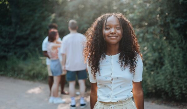 A teenage girl smiling confidently outdoors on a sunny day with friends in the background, representing presence and connection.
