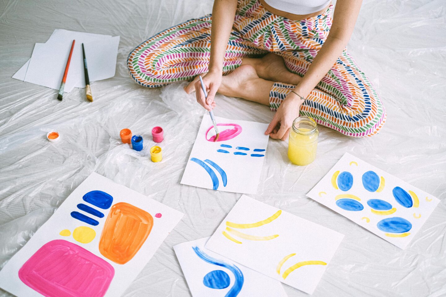 A person sitting cross-legged on the floor painting colorful abstract shapes on paper, surrounded by vibrant paint pots and brushes.