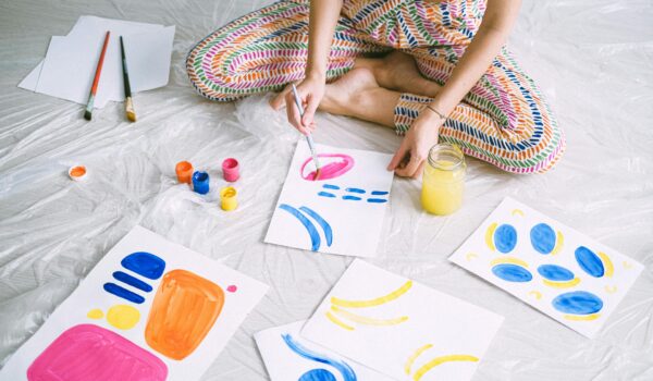 A person sitting cross-legged on the floor painting colorful abstract shapes on paper, surrounded by vibrant paint pots and brushes.