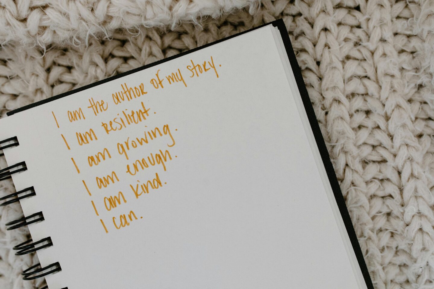 A spiral notebook with handwritten affirmations in gold ink reading 