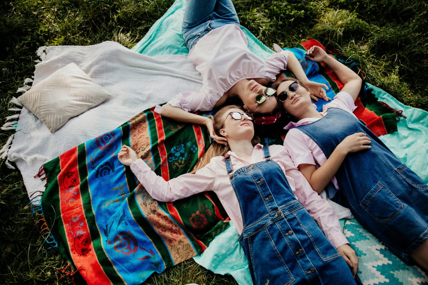 Three teenage girls lying together on colorful blankets in the grass, smiling and relaxing outdoors on a sunny day.