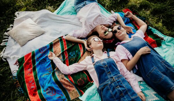 Three teenage girls lying together on colorful blankets in the grass, smiling and relaxing outdoors on a sunny day.