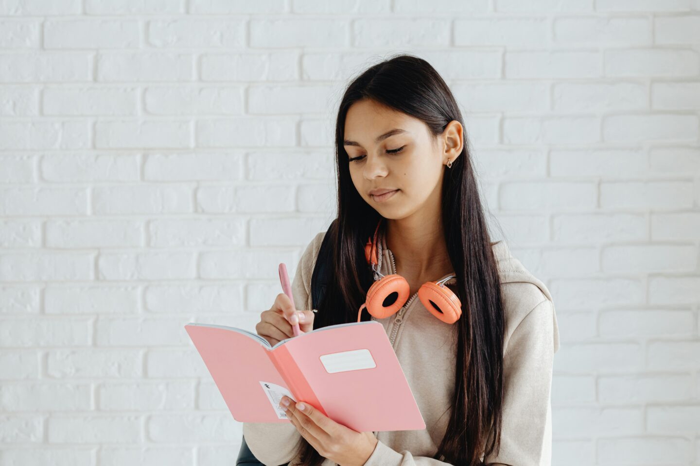A smiling teenage girl with headphones around her neck writing in a pink journal, representing journaling for teen mental health.