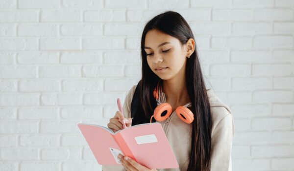 A smiling teenage girl with headphones around her neck writing in a pink journal, representing journaling for teen mental health.