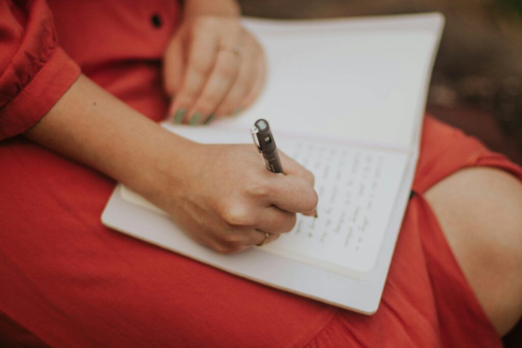 A close-up of a person in a red outfit writing in an open journal on their lap, symbolizing a daily journaling practice for mental wellness.