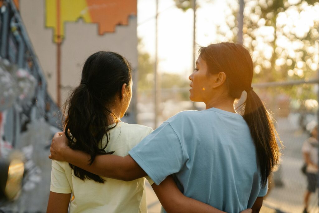 Two young women walking together outside at sunset, one with her arm around the other in a gesture of support and friendship.