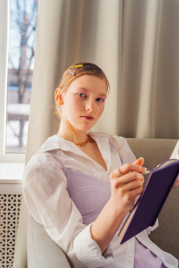 A teenage girl with colorful face gems writing in a journal while sitting by a bright window, reflecting on her thoughts.