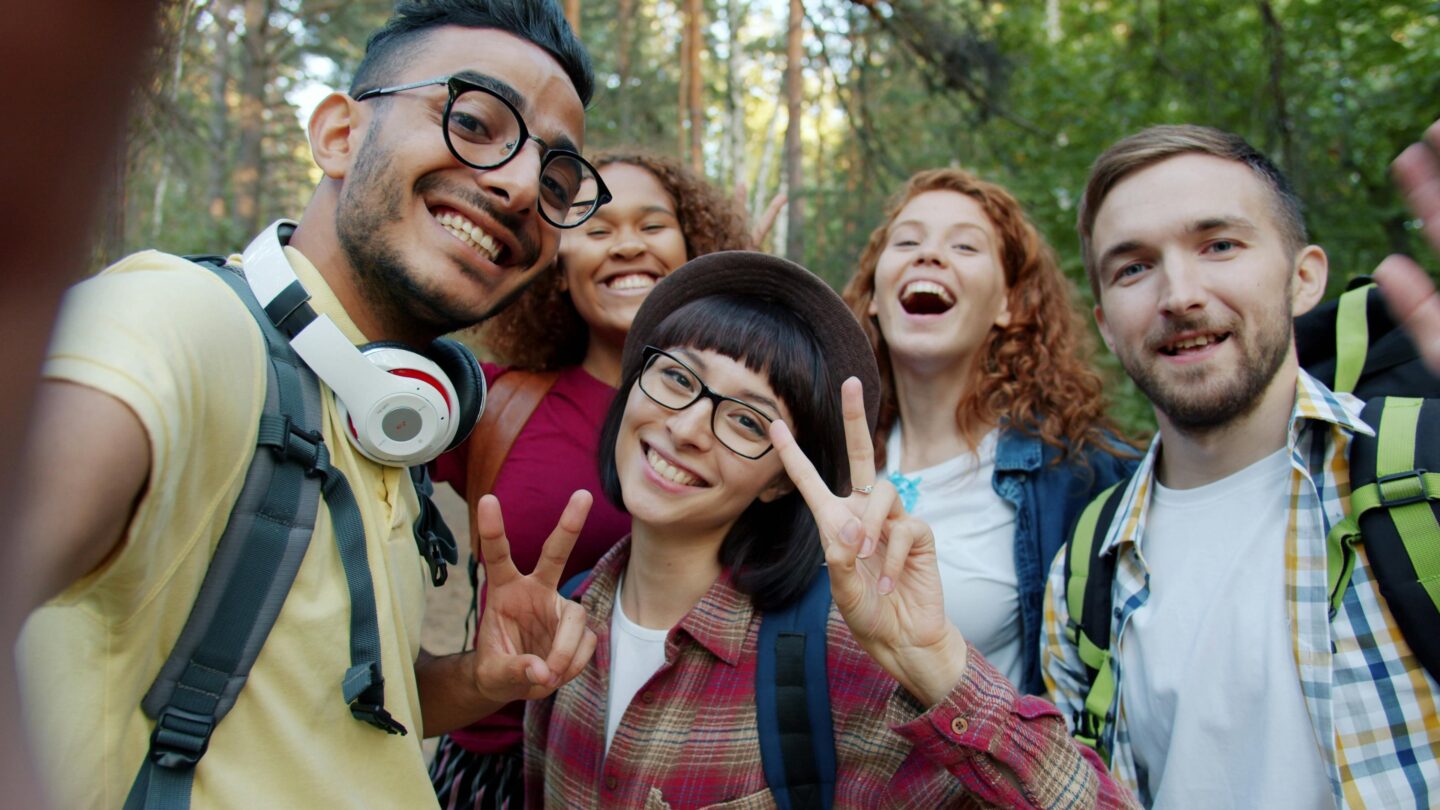 A diverse group of five young adults smiling and taking a selfie together outdoors in a forest, wearing backpacks and showing peace signs