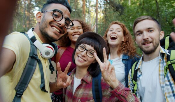 A diverse group of five young adults smiling and taking a selfie together outdoors in a forest, wearing backpacks and showing peace signs