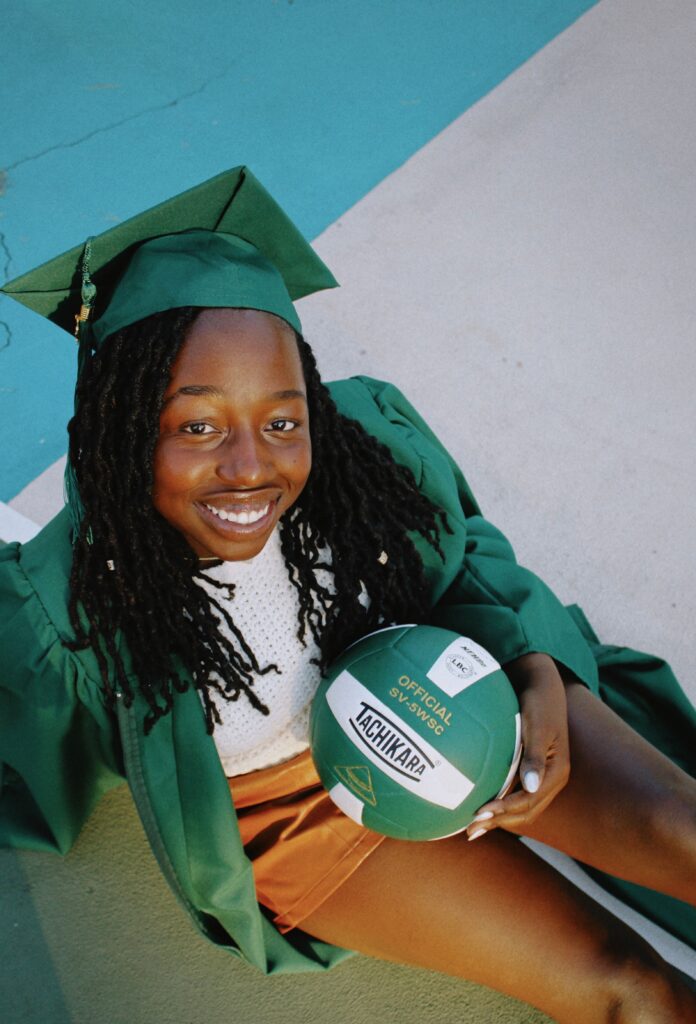 A smiling young graduate in a green cap and gown sits outdoors holding a green and white volleyball, celebrating both her academic and athletic achievements.