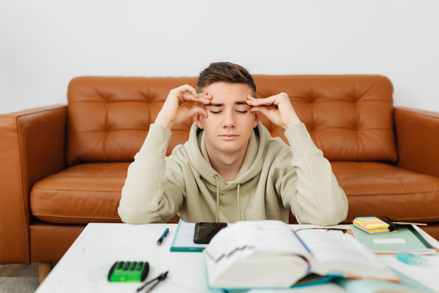 A high school student feeling academic pressure, eyes closed and hands on his temples while studying at home.