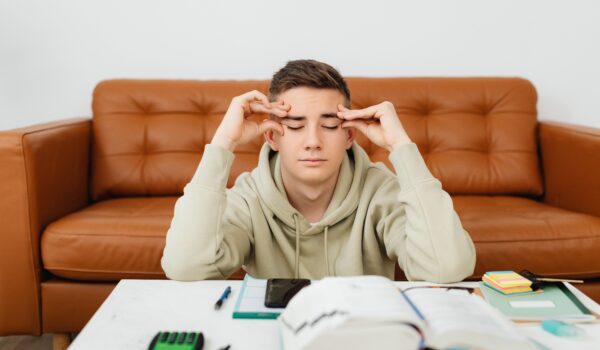 A high school student feeling academic pressure, eyes closed and hands on his temples while studying at home.