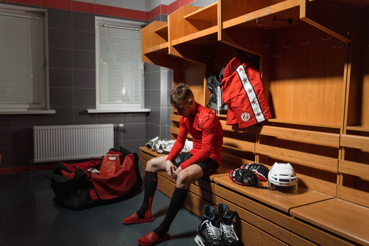 A young hockey player in a red jersey sits alone on a locker room bench, head down and looking pensive, surrounded by his gear, skates, helmet, and gloves.