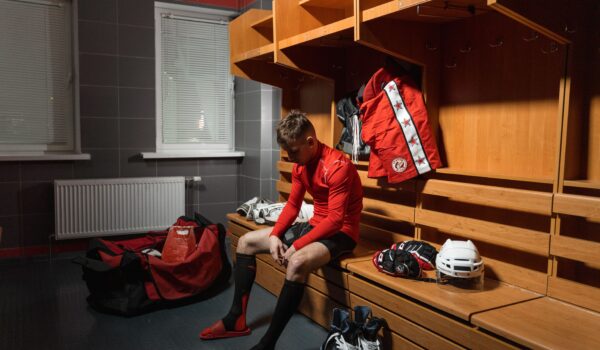 A young hockey player in a red jersey sits alone on a locker room bench, head down and looking pensive, surrounded by his gear, skates, helmet, and gloves.