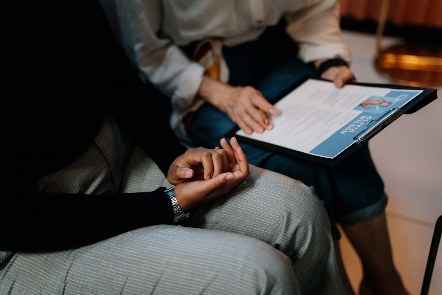 Close-up of clasped hands in a therapy setting, with a clinician's clipboard and notes visible in the background.