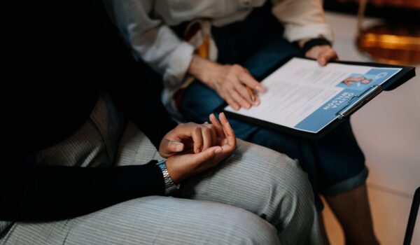 Close-up of clasped hands in a therapy setting, with a clinician's clipboard and notes visible in the background.