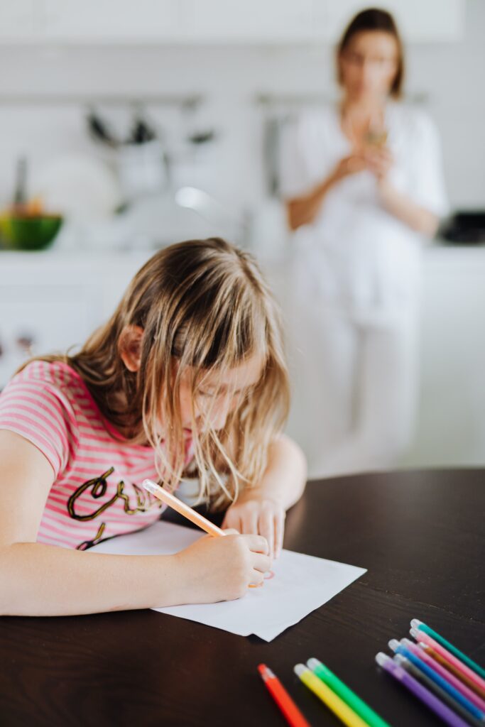 Young girl focused on writing at a kitchen table surrounded by colored pencils, with a parent watching from a distance in the background.