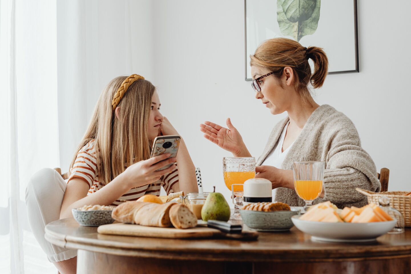 A parent and teenager sitting across from each other at a kitchen table in a calm but serious conversation, both looking at each other.