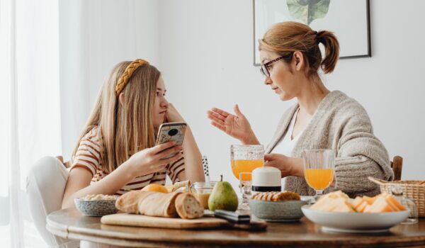 A parent and teenager sitting across from each other at a kitchen table in a calm but serious conversation, both looking at each other.