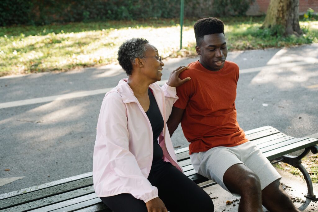 Teen boy sitting on a park bench, talking with his mom who has a supportive hand on his shoulder, both smiling in an easy, open conversation.
