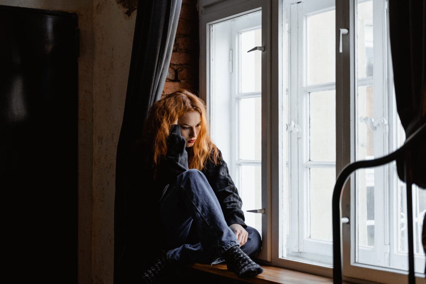 A young woman sits curled up alone by a window, looking down with a distant, weighed-down expression.