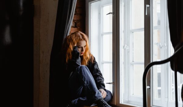 A young woman sits curled up alone by a window, looking down with a distant, weighed-down expression.