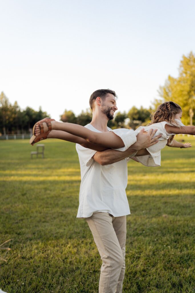 Father playfully swinging his young daughter through the air, both smiling, in a sunny open field.